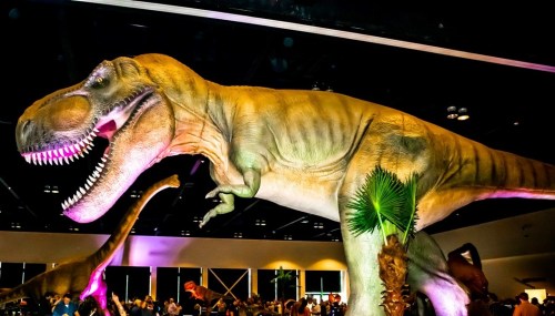 A large, lifelike T. rex dinosaur model in a crowded exhibition space, with palm plants in the foreground.