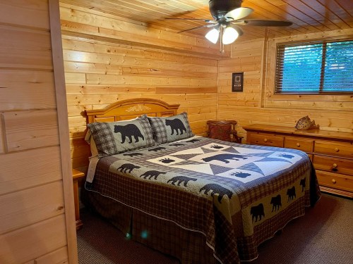 Cozy wooden bedroom featuring a bed with a bear-patterned quilt, dresser, and ceiling fan. Natural light from a window.