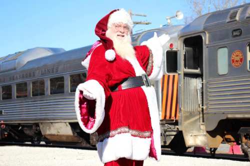 Santa Claus in a red suit stands beside a vintage train, smiling and waving in a festive outdoor setting.