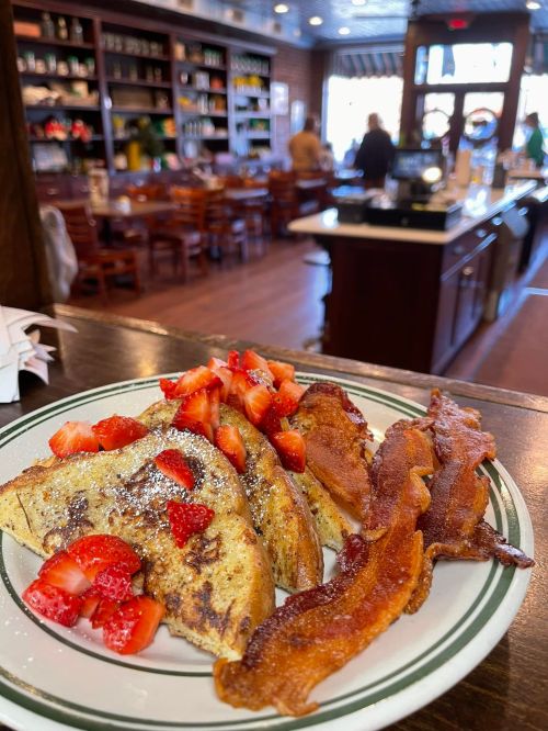 A plate of French toast topped with strawberries and crispy bacon, set in a cozy restaurant interior.