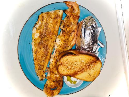 A blue plate with seasoned fish fillets, shrimp, a baked potato wrapped in foil, and a slice of toasted bread.