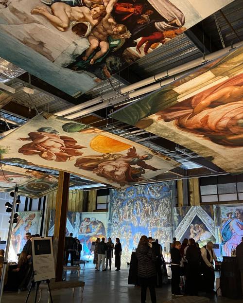 A gallery with large, hanging reproductions of Michelangelo's artwork, featuring visitors admiring the displays.