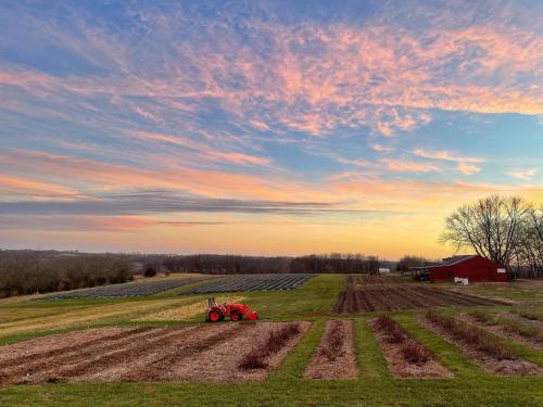 A vibrant sunset over a farm with fields, a red barn, and a tractor in the foreground.