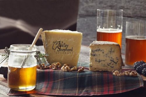 A wooden platter with two cheese varieties, a jar of honey, walnuts, and two glasses of amber beer on a plaid cloth.