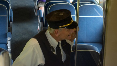 A train conductor in uniform leans against a pole inside a train car, with blue seats visible in the background.