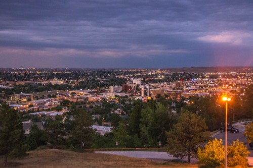 A panoramic view of a city at dusk, with lights twinkling and a cloudy sky overhead. Trees frame the foreground.