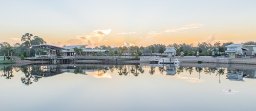 A serene lake at sunset, reflecting trees and buildings, with a peaceful sky and soft clouds above.