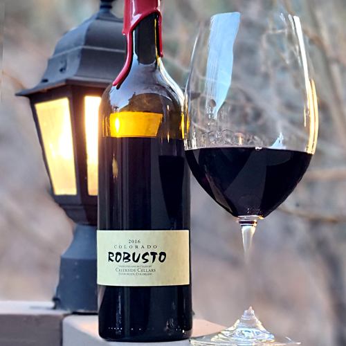 A bottle of 2016 Colorado Robusto wine next to a glass filled with red wine, illuminated by a lantern.
