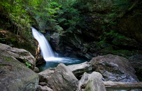 A serene waterfall cascades over rocks into a tranquil pool, surrounded by lush green foliage and rugged terrain.