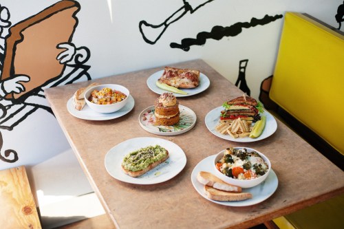 A table with various colorful dishes, including salads, toast, and baked goods, against a playful mural backdrop.