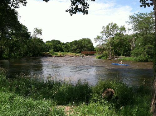 A serene river scene with a kayaker paddling, surrounded by lush greenery and trees under a partly cloudy sky.