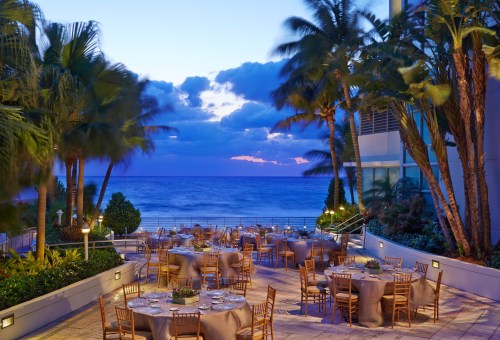 Outdoor dining area with round tables set up, surrounded by palm trees and overlooking the ocean at sunset.