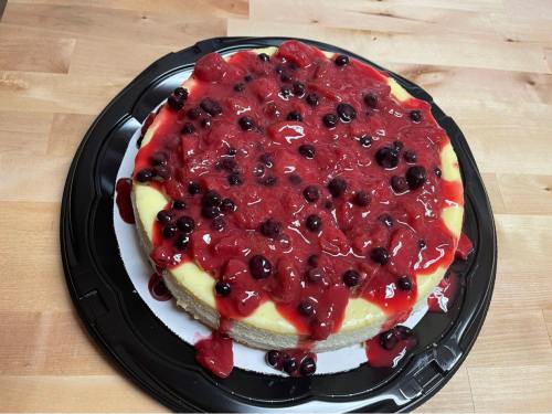 A cheesecake topped with a glossy red berry sauce and blackberries, placed on a black plastic tray.
