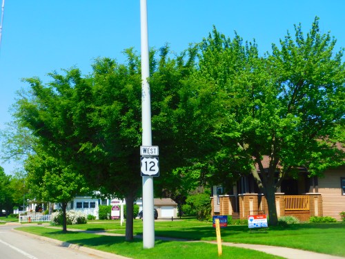 A clear blue sky over a street lined with green trees and houses, featuring a sign for Route 12 West.