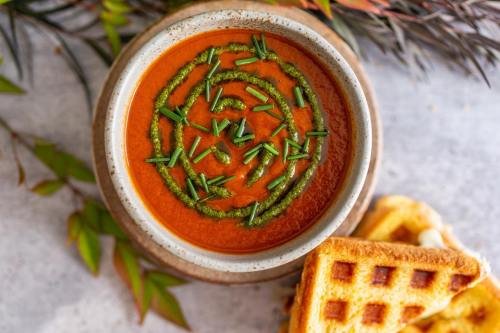 A bowl of tomato soup topped with green swirl and chopped herbs, accompanied by a piece of toasted bread.