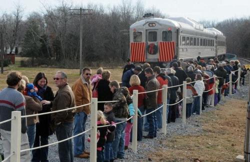 A long line of people waiting near a vintage train on a rural track, with trees in the background.