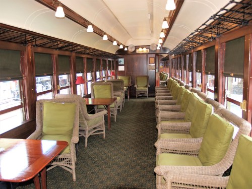 Interior of a vintage train car featuring green cushioned chairs and wooden tables, with large windows and warm lighting.