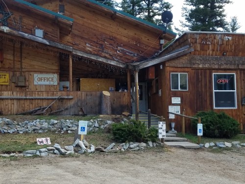 A rustic wooden building with an "Office" sign and an "Open" sign, surrounded by trees and gravel pathways.