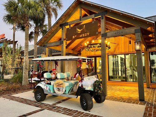 A festive golf cart decorated for the holidays is parked outside a brewery with a wooden entrance and palm trees.