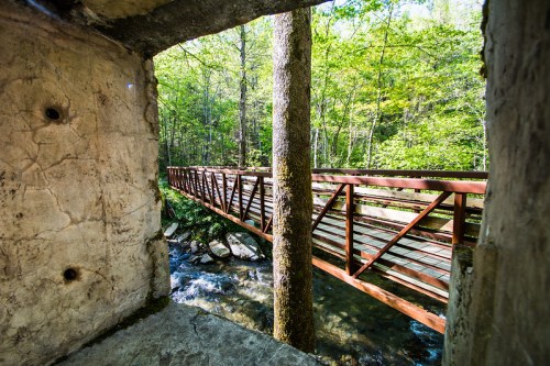 View of a wooden bridge crossing a stream, framed by a stone wall and surrounded by lush green trees.