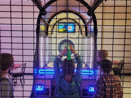 A group of four people stands in front of a game machine, preparing to play, with colorful lights and a scoreboard visible.