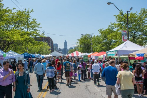A bustling street fair with colorful tents, people walking, and city buildings in the background under a clear blue sky.