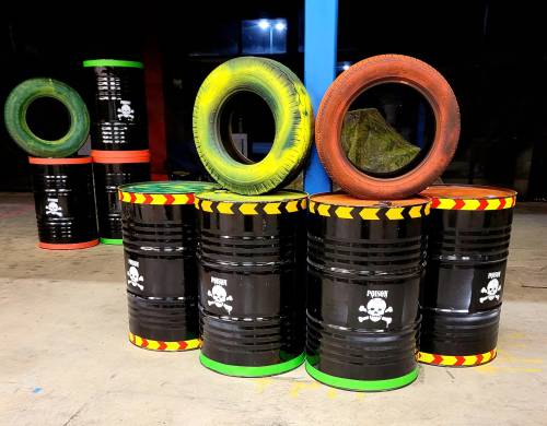 A stack of black barrels with hazard symbols, surrounded by colorful tires, in a dimly lit indoor space.