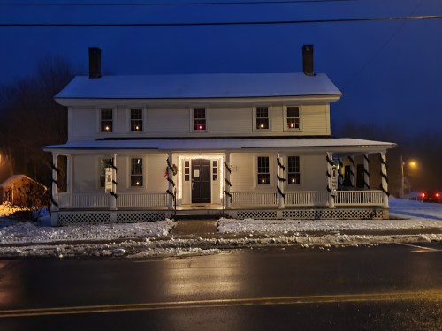 A two-story white house with a porch, snow-covered ground, and warm lights glowing in the windows at dusk.