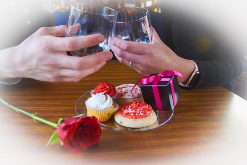 Two hands clink glasses over a plate of decorated cookies and a small gift box, with a red rose nearby.