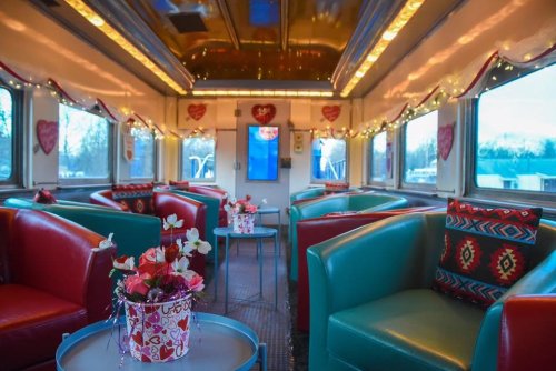 A colorful train car interior decorated with hearts and lights, featuring cozy seating and floral centerpieces.