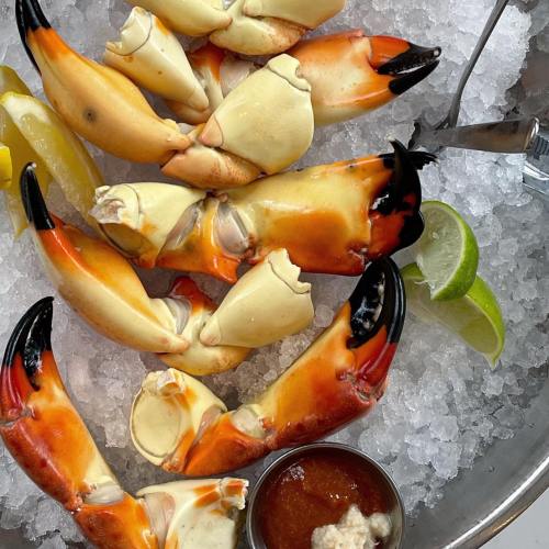 A platter of crab claws on ice, garnished with lemon and lime wedges, accompanied by a small bowl of dipping sauce.