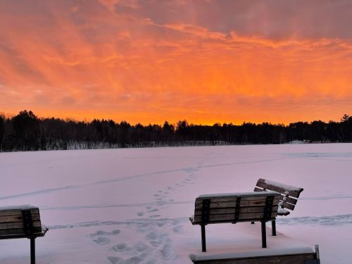 A serene winter scene with two benches overlooking a snowy landscape and a vibrant orange sunset in the sky.