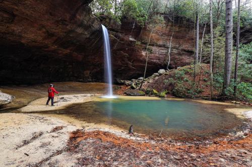 A person in a red jacket stands by a serene waterfall and pool surrounded by lush greenery and rocky cliffs.