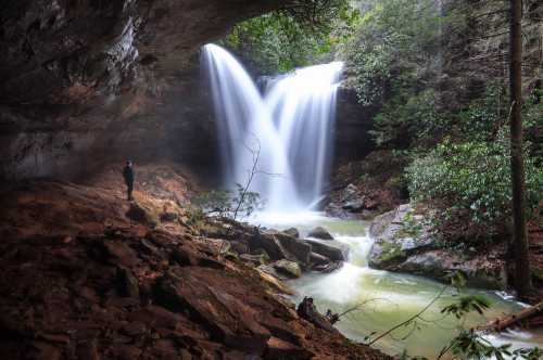 A serene waterfall cascades into a rocky pool, surrounded by lush greenery and a person standing nearby.