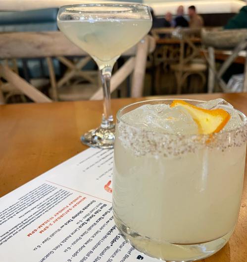 Two cocktails on a wooden table: a glass with a salted rim and orange garnish, and a martini in the background.