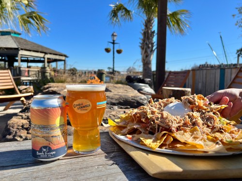 A plate of nachos topped with pulled pork and a dollop of sour cream, alongside a can and glass of beer, outdoors.