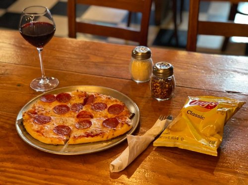 A plate of pepperoni pizza on a wooden table, accompanied by a glass of red wine and a bag of Lay's chips.