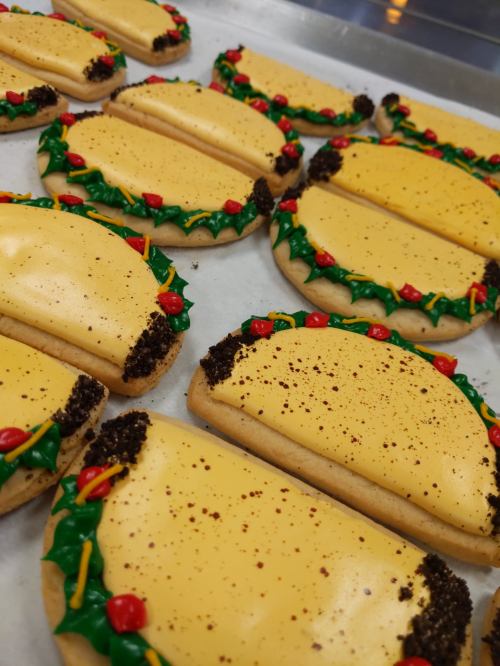 Colorful cookies shaped like tacos, decorated with yellow icing, green frosting, and red candy toppings.