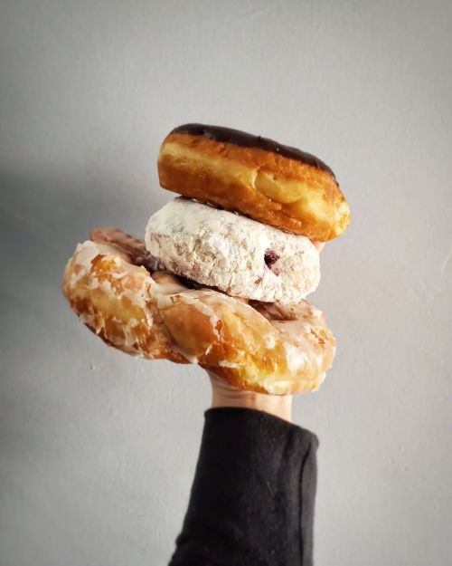 A hand holding three stacked donuts: a chocolate-covered, a powdered, and a glazed donut against a gray background.