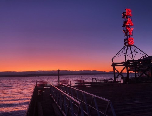 A vibrant sunset over water with a neon sign reading "RAYS" on a pier, casting a warm glow against the evening sky.