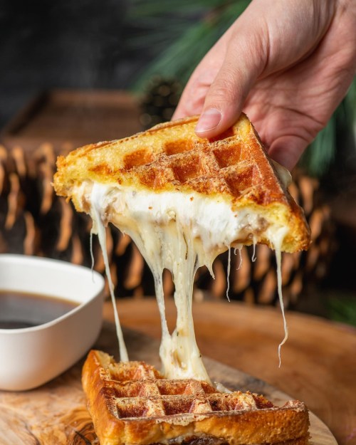 A hand holds a cheesy waffle, with melted cheese stretching, next to a small cup of coffee on a wooden surface.
