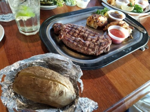 A grilled steak on a metal plate with dipping sauces, alongside a foil-wrapped baked potato on a wooden table.