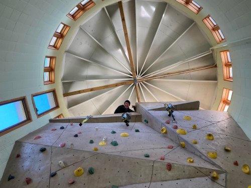 A person climbs a rock wall inside a circular building with a large, spiraled ceiling above.