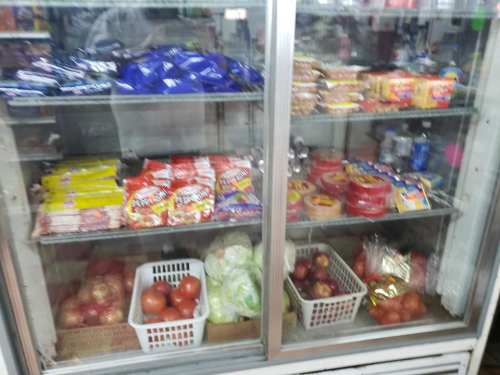 A glass-front refrigerator filled with snacks, drinks, and fresh produce like apples and lettuce.