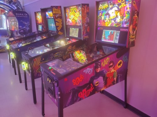 Three colorful pinball machines featuring comic book themes, including Batman and Robin, in a vibrant arcade setting.