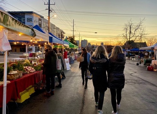 Two people walk through a bustling farmers market at sunset, surrounded by colorful tents and fresh produce.