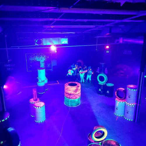 A vibrant indoor laser tag arena with neon lights, barrels, and players in action amidst a colorful atmosphere.