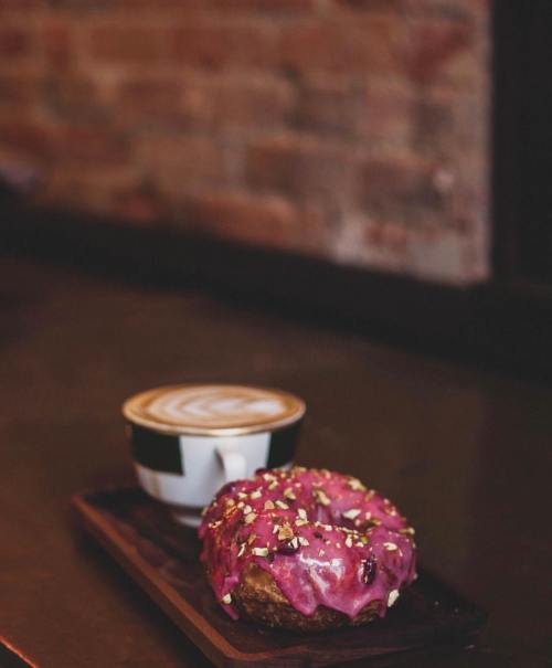 A pink frosted donut with pistachios sits on a wooden tray next to a latte with a decorative foam design.