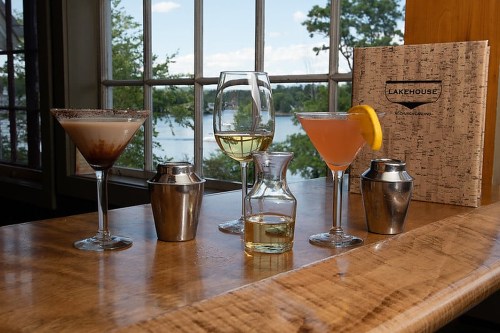 A variety of cocktails on a wooden bar with a scenic lake view through large windows.