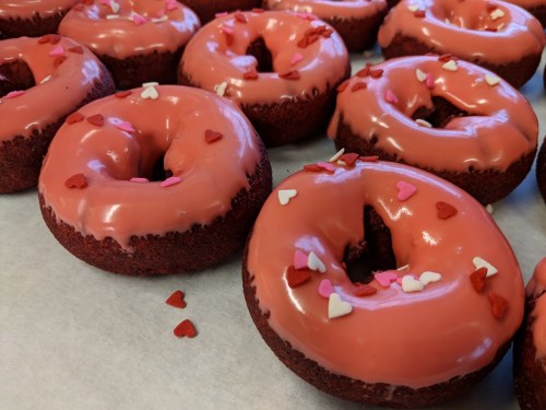 A close-up of red velvet donuts topped with pink icing and heart-shaped sprinkles on a white surface.